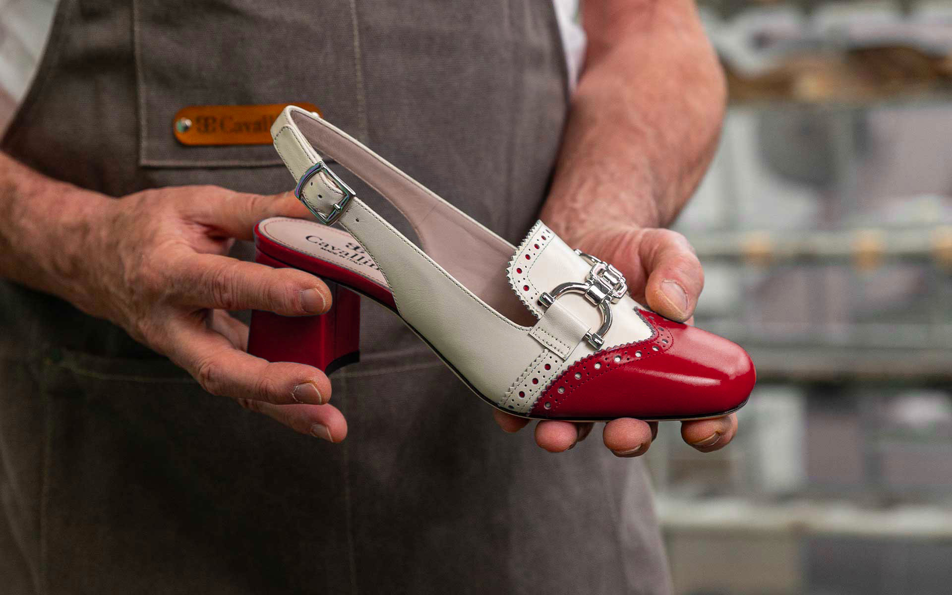 Maestro artigiano Cavallini tiene in mano una scarpa Donna in pelle bicolore con tacco rivestito e fibbia decorativa, nel laboratorio di Santa Maria a Monte